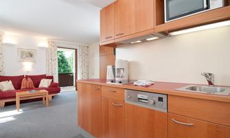 A modern kitchenette with wooden cabinets and a microwave. In the background, there is a cozy living area with a red sofa.