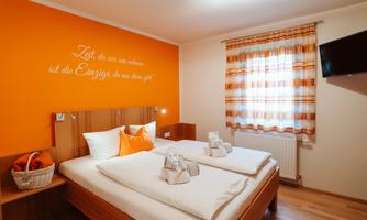 A cozy bedroom with a double bed, an orange wall paint, and a television. The window decoration and towels create an inviting atmosphere.