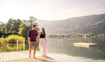 A couple walks along the water, holding hands. In the background, there are mountains and a calm lake.
