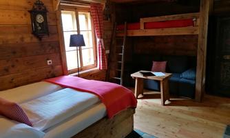 A cozy room in rustic style with a double bed and a loft bed. There are wooden paneling, a small table, and a window with a view outside.
