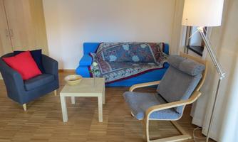 A cozy living room with a blue sofa, a gray armchair, and a small table. A floor lamp and a flower vase complement the decor.