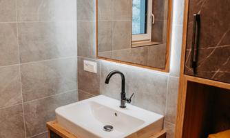 A modern bathroom with an elegant sink unit and a large mirror. The walls are tiled with gray tiles and a small window provides natural light.