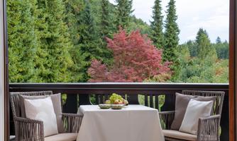 A cozy balcony with two chairs and a table. View of lush, green trees and a blooming red tree in the background.