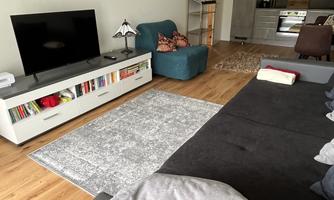 A modern living room with a large sofa, a television, and a table. The room has a warm wooden floor and a cozy atmosphere.