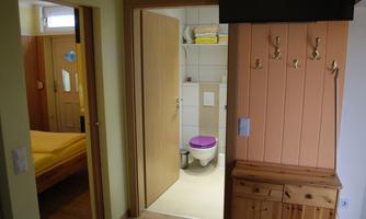 A small hallway with wooden paneling and a toilet in the background. On the left side, a bedroom with a bed is visible.