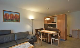 A modern living room with a black sofa and a wooden dining table. A picture of tulips hangs on the wall, and the kitchen has an open design.