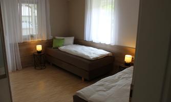 A bright room with two beds, one on a wooden platform and one with a pillow and a blanket. Two table lamps and large windows let in plenty of light.