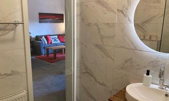 A modern bathroom with marble walls and a round mirror. In the background, there is a cozy living area with a gray sofa and colorful cushions.