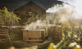 A wooden bath with steaming water in an idyllic setting. In the background, a wooden house and nature can be seen.