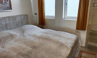 A bedroom with a large bed and a bright ceiling. The walls are painted light and there are two windows with curtains.
