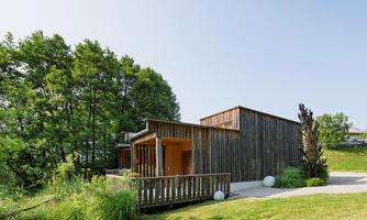 Modern wooden houses by the shore of a tranquil lake, surrounded by lush nature. In the background, picturesque trees and a blue sky can be seen.