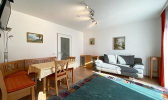 A bright living room with a cozy seating area and a dining table. The walls are decorated with pictures and the light is inviting.