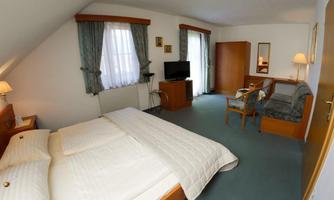 A bright hotel room with a double bed and a cozy seating area. There are windows with curtains and a television in the room.