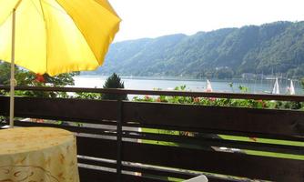 A beautiful view of a lake with boats and green hills in the background. A round table with a yellow sunshade is on the terrace.