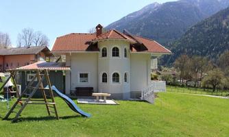 A beautiful house with a red roof and large windows, surrounded by green grass. In the foreground, there is a playground structure with a slide and a swing.