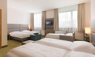 A modern hotel room with two bed areas and an additional sofa bed. Large windows provide plenty of natural light.