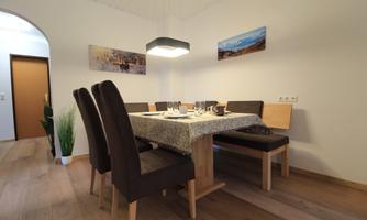 A modern dining area with a wooden table and comfortable chairs. The walls are white, and there are pictures hanging with landscape motifs.