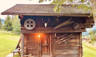 A rustic wooden cabin with traditional decorations and tools. The building is situated in a green landscape with trees and mountains in the background.