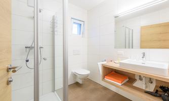 A modern bathroom with a shower and a sink. The walls are light-tiled, and there is a wooden shelf with towels.