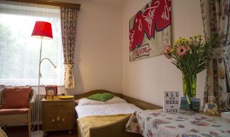 A cozy room with a single bed and a colorful lampshade. The table is decorated with a flower vase and a sign that says "ALL YOU NEED IS LOVE."