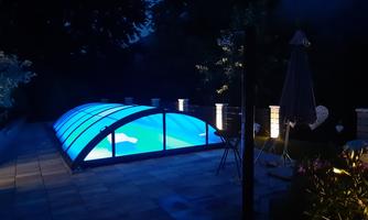 A illuminated pool under a transparent pool roof at night. The area is surrounded by plants and a sunshade, framed by gentle lighting.