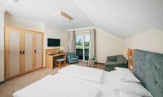 A modern hotel room with two single beds and a cozy seating area. There is a television and access to a balcony with an outdoor view.