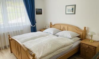 A cozy bedroom with a wooden bed and white comforters. The room has large windows with blue curtains and a bedside lamp.