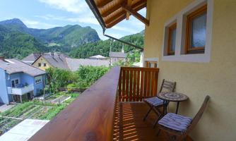 A balcony with a wooden railing and two chairs overlooks green mountains and a village. The surroundings are quiet and idyllic.