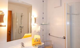 A modern bathroom with a sink, a mirror, and a shower. The floor is tiled in blue, and there is a wooden shelf.
