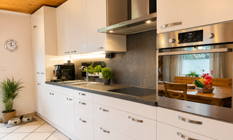 A modern kitchen with white cabinets and a dark countertop. In the background, a dining area with a table and plants is visible.