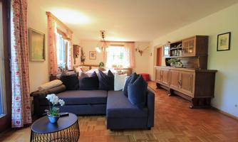 A cozy living room with a large sofa and a small table. The windows let in a lot of light, and there are decorative elements as well as a wooden furniture cabinet.