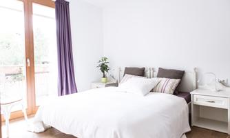 A bright bedroom with a large bed and white bedding. There is a desk, a chair, and a plant next to the window.