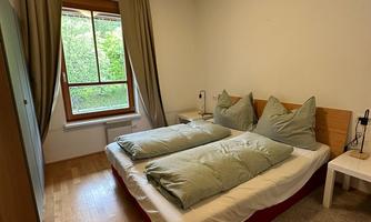 A simple bedroom with a double bed and green cushions. The room has a view of the outdoors and a warm wooden floor.
