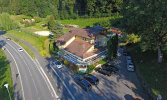A restaurant on a street, surrounded by green meadows and trees. In front of the building, several cars are parked.