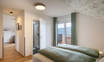 A cozy bedroom with two beds and a large window. The room is modernly furnished and has access to a balcony with a view outside.