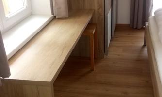 A simple, well-lit room with a desk and a chair. The floor is made of wood, and the walls are paneled with wood.