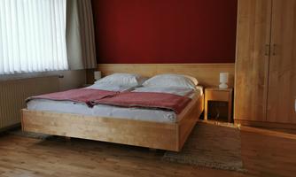 A cozy bedroom with a wooden bed and red walls. The room is brightly lit and has a simple bedside table.