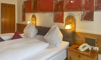 A cozy bedroom with two beds and white pillows. The walls are decorated with red ceramic tiles and a bedside lamp glows softly.