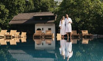 A couple in bathrobes stands at the edge of a swimming pool. In the background, there are loungers and a beautiful, green environment visible.