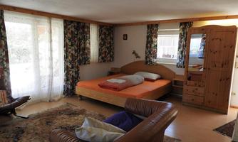 A cozy bedroom with a large bed and a wooden dresser. The windows are decorated with colorful curtains and there is a comfortable armchair in the room.