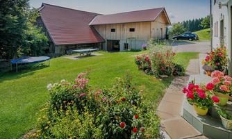 A beautiful garden with colorful flowers and a well-kept lawn. In the background, rustic buildings and a car can be seen.