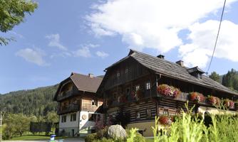 A traditional wooden house with colorful flowers at the windows. The surroundings are green and picturesque, with a clear blue sky.