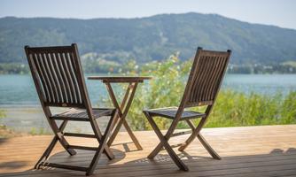 Two wooden lounge chairs are positioned at a table on a wooden terrace. In the background, a beautiful lake and green mountains can be seen.