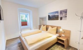A bright guest room with a double bed and two large pillows. Photos of mountains hang on the wall, and there is a window overlooking the outside.