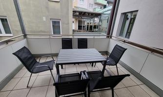 A small balcony with a table and six chairs. The walls are light, and there is a view of neighboring buildings.