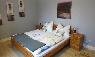 A cozy bedroom with a double bed and light wooden furniture. The walls are painted in soft gray tones, and some pictures adorn the room.