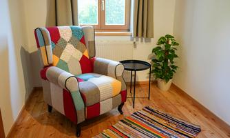 A colorful armchair stands beside a small table in a bright room. A striped carpet lies on the floor and a plant stands in the corner.