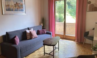 A cozy living room with a gray sofa and pink cushions. Large windows provide plenty of daylight and a view of the greenery.