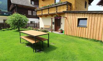 A cozy garden with a wooden house and a seating area made of tables and benches. The green grass and the trees give the surroundings an inviting atmosphere.
