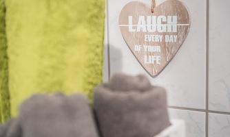 A dark towel and a green bath towel are visibly placed on a shelf. In the background, there is a heart-shaped sign that reads "Laugh every day of your life."